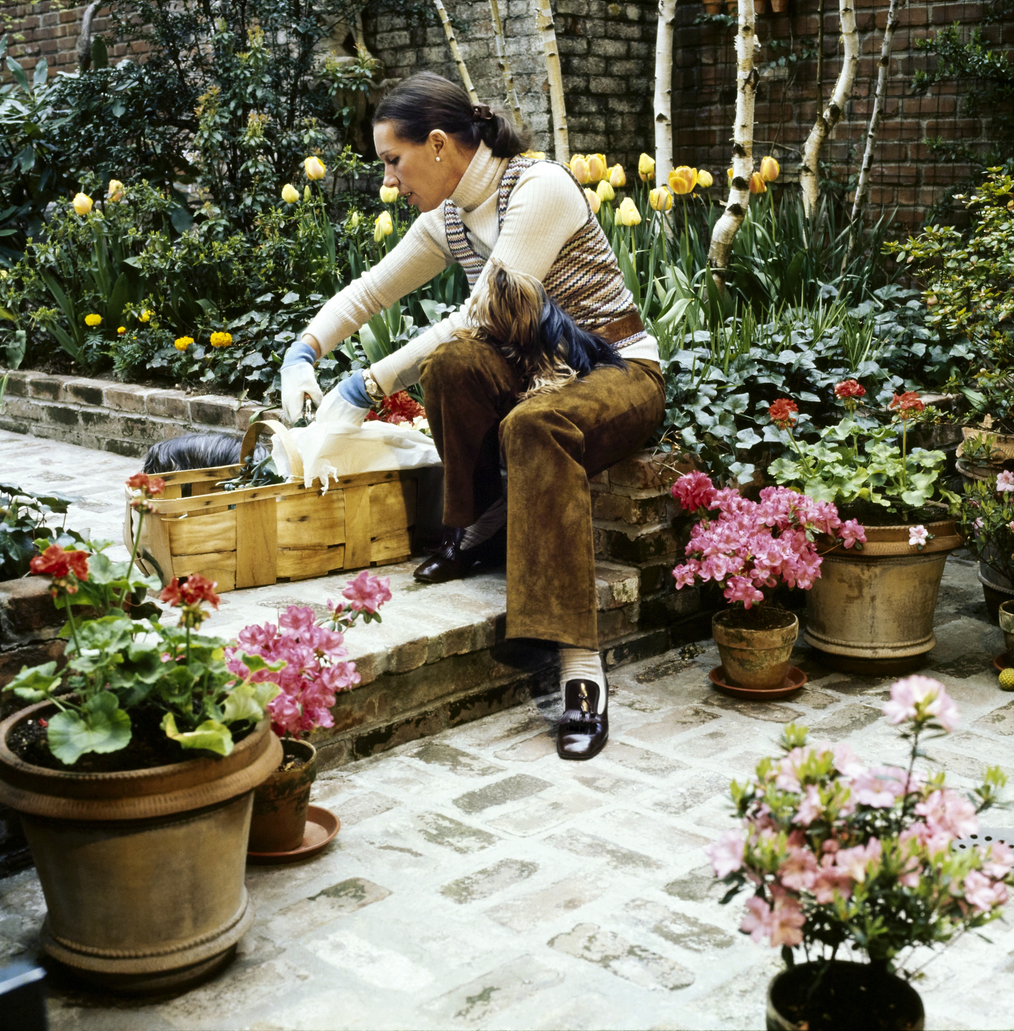 At the New York City home of Françoise and Oscar de la Renta, Françoise, wearing brown suede pants and a turtleneck, sits on a bench cutting flowers in her garden