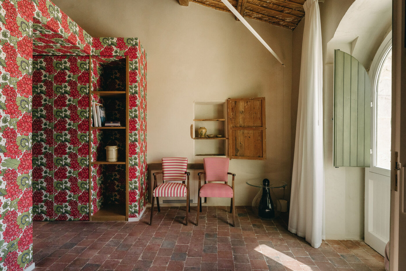 A Mediterranean farmhouse has been transformed into a boutique hotel mixing rustic tiled flooring with wooden ceilings and shelves, and red, green, and white wallpaper floral motifs.