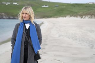 Ashley Jensen as DI Ruth Calder in Shetland. She is standing on the beach wearing a warm grey cashmere coat, with an electric blue scarf wrapped around her neck with the ends flapping in the end. Ruth's body is facing the camera but her face is looking off to the side, out to sea