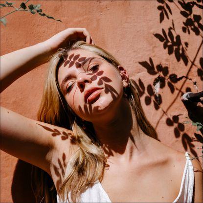 woman with blonde hair leaning against a wall with shadows of leaves on her face