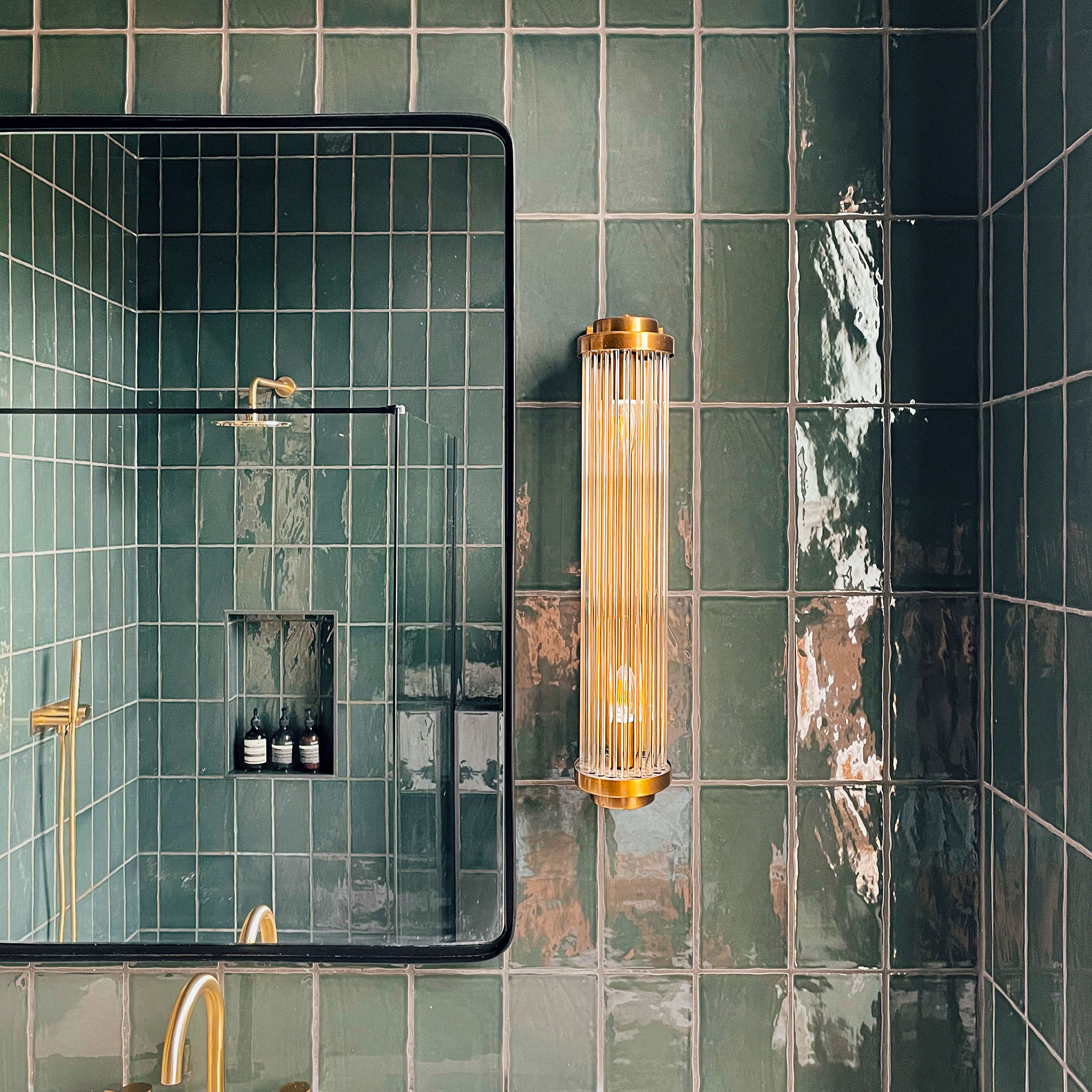 Dark green tiled bathroom with two glass and gold wall light beside mirror reflecting shower area