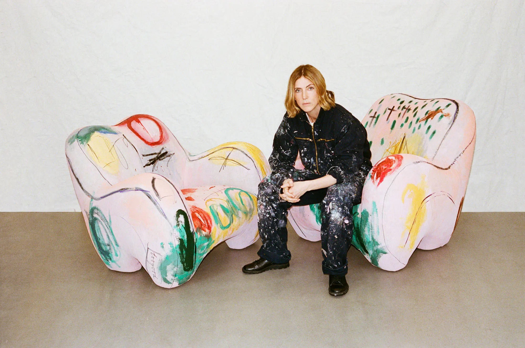 A young woman dressed in a black, covered-in-paint boiler suit sits atop one of two whimsically shaped armchairs covered in bold, abstract green, black, red, and yellow brushstrokes.
