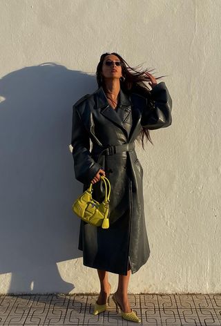 A woman wearing an oversize leather trench coat as a dress with a green bag and green pumps.