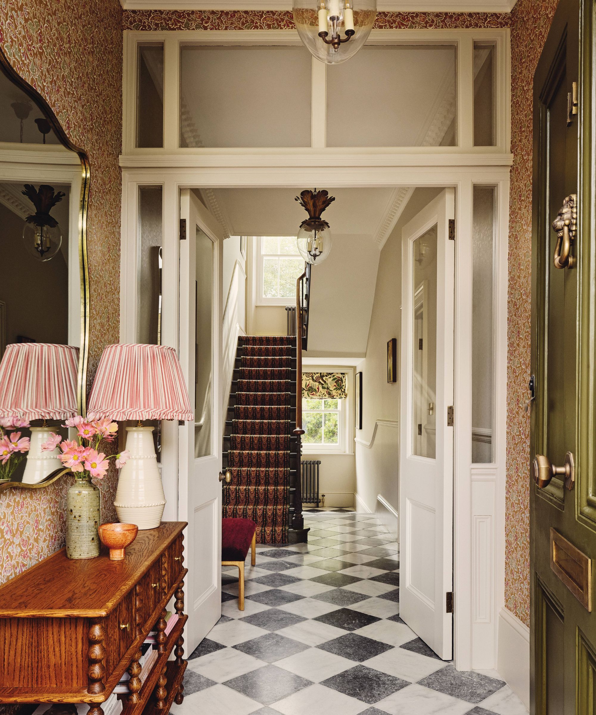 A traditional hallway with a black-and-white checkered marble floor. The is a wooden side table on which is a pink lamp and a gold mirror hangs above.