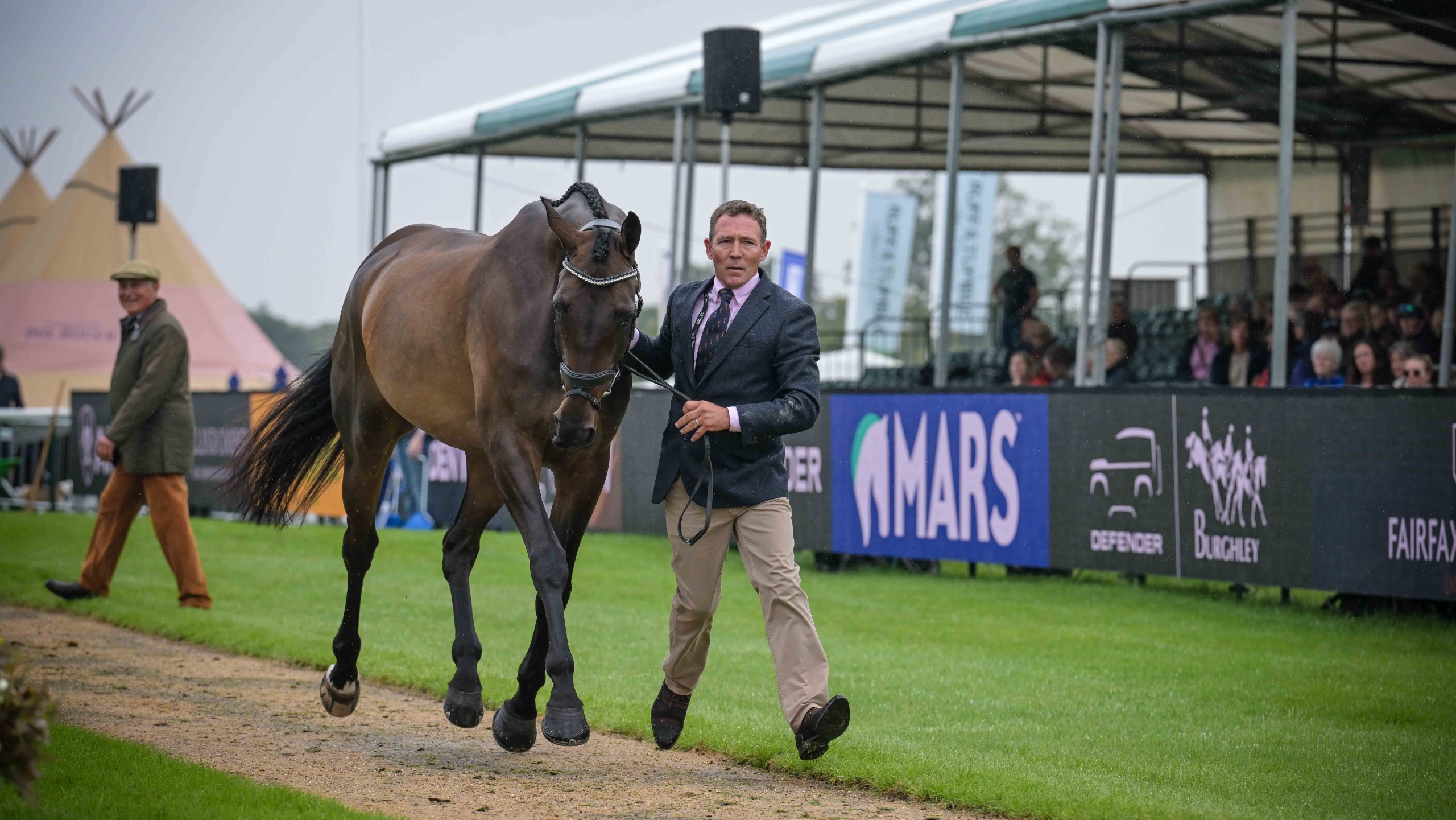 Austin O'Connor, a man wearing a blue jacket, a pink shirt and a tie, trots up a bay horse called Sixmilewater at Burghley 2026