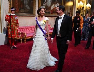 Kate Middleton wearing a white dress and diamond tiara walking next to a man in a tuxedo with a palace guard standing behind her