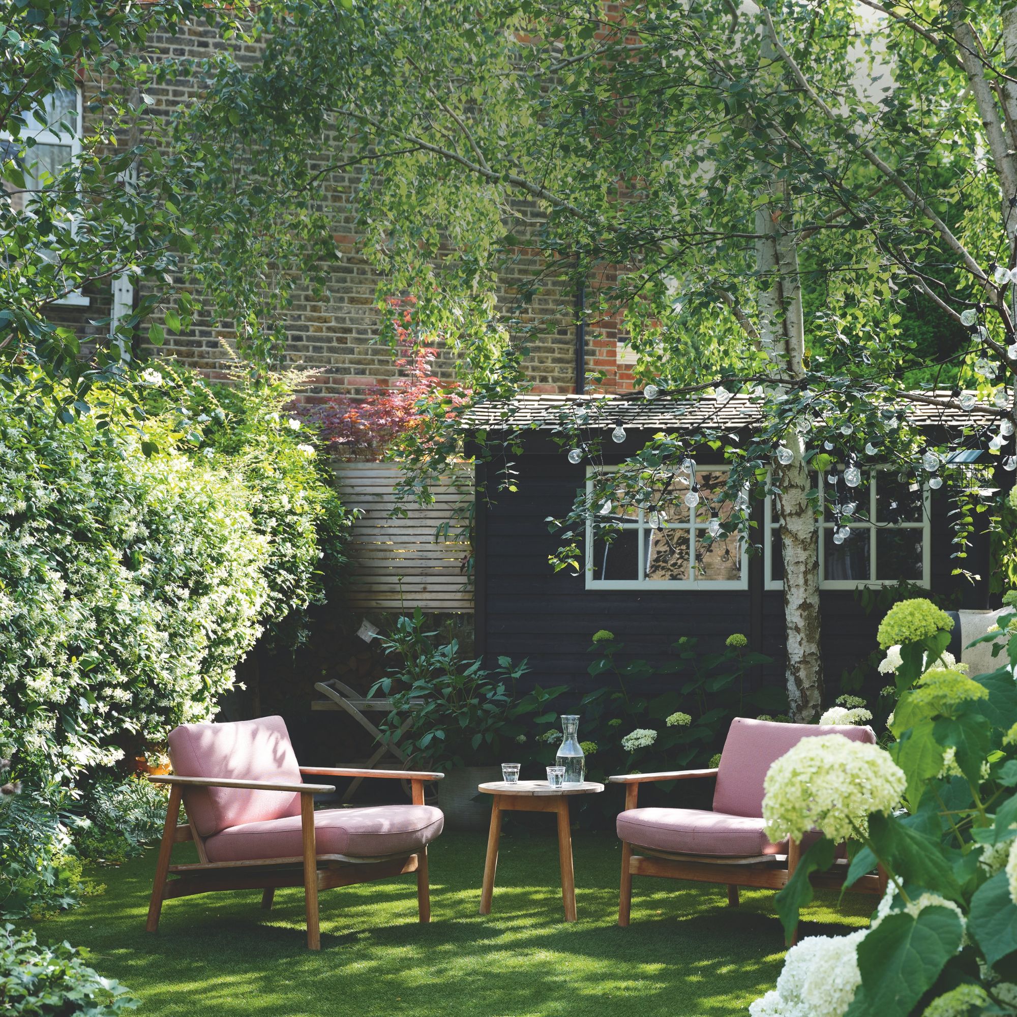 A black outbuilding in a garden with a tree and seating in front of it