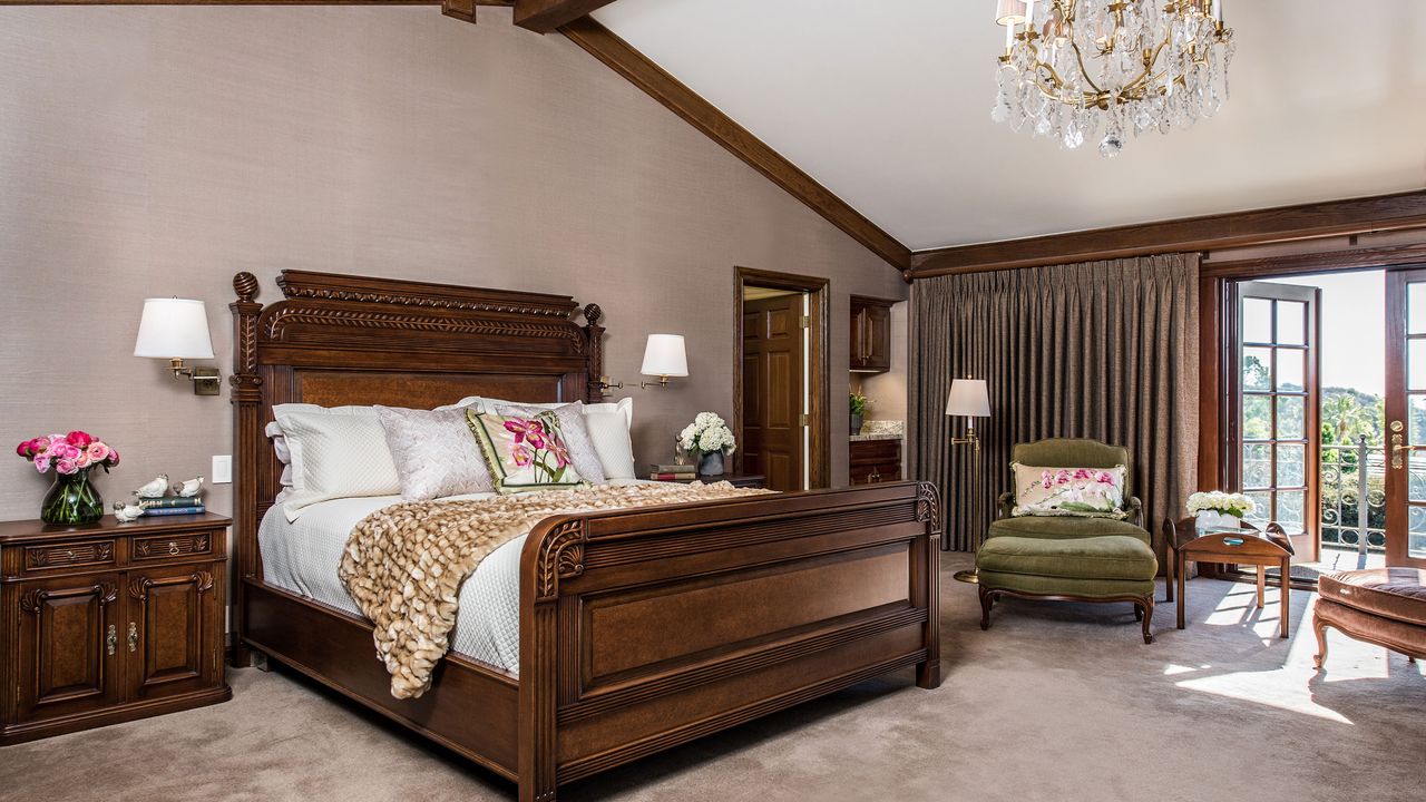 Large antique bed in mahogany with silk bedsheets and vase of pink roses on bedside table and green chair