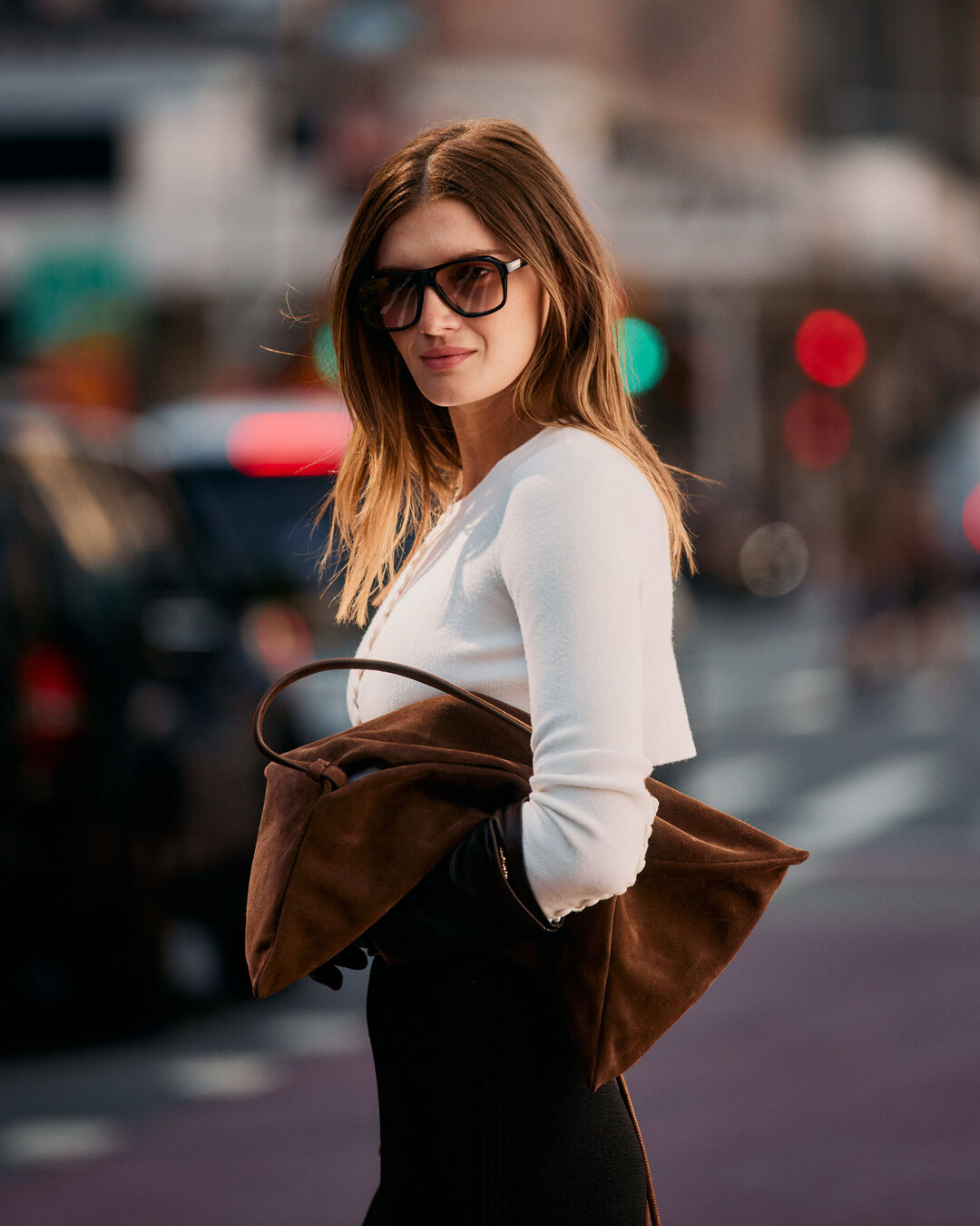 Woman carries brown suede bag while wearing a white cardigan and black skirt.