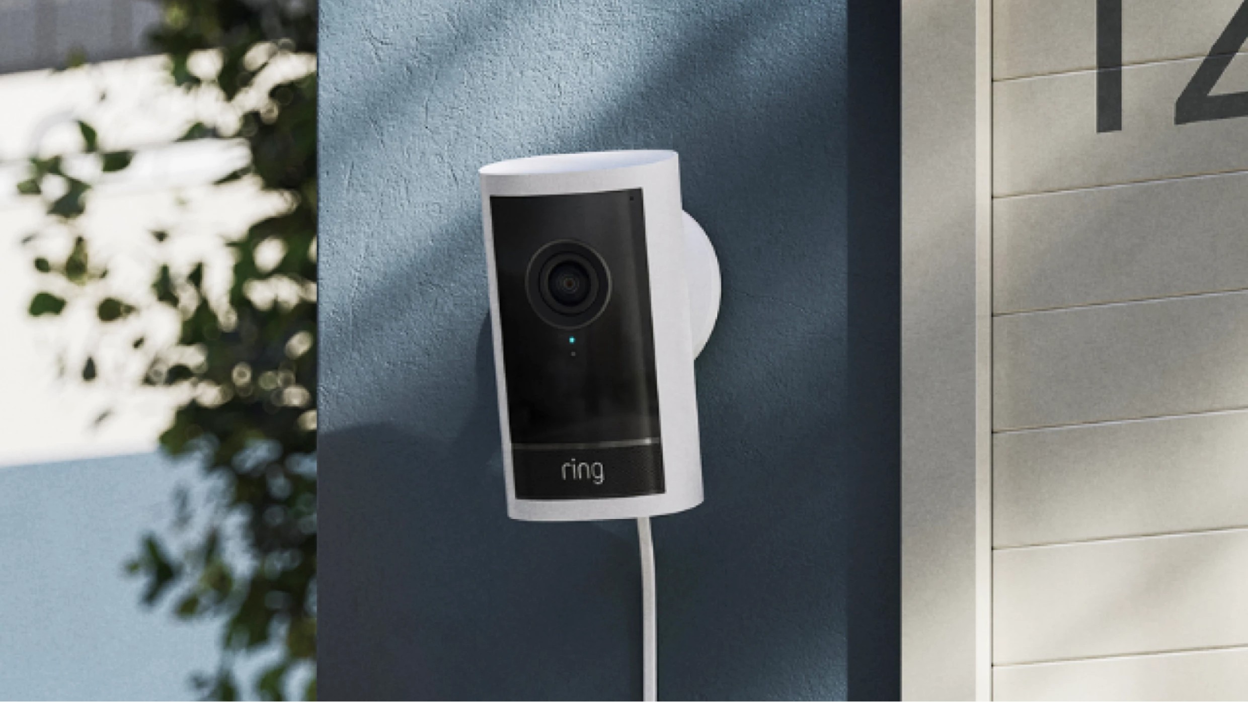 A white and black Ring security camera, connected by a white cable, is mounted on a dark blue exterior wall next to a light-colored wall with the number &#039;12&#039;.