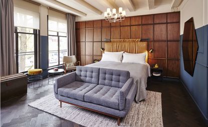 The Hoxton bedroom with wood panelling behind bed and grey sofa at foot. Grey herringbone floor tiles and hexagonal mirror on blue and white wall