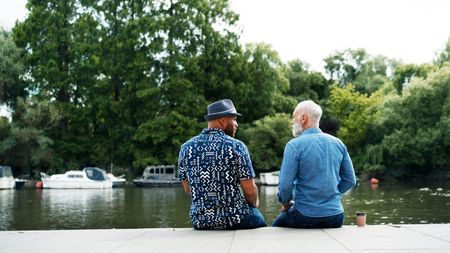 Two older men sit on the side of a lake and talk. 