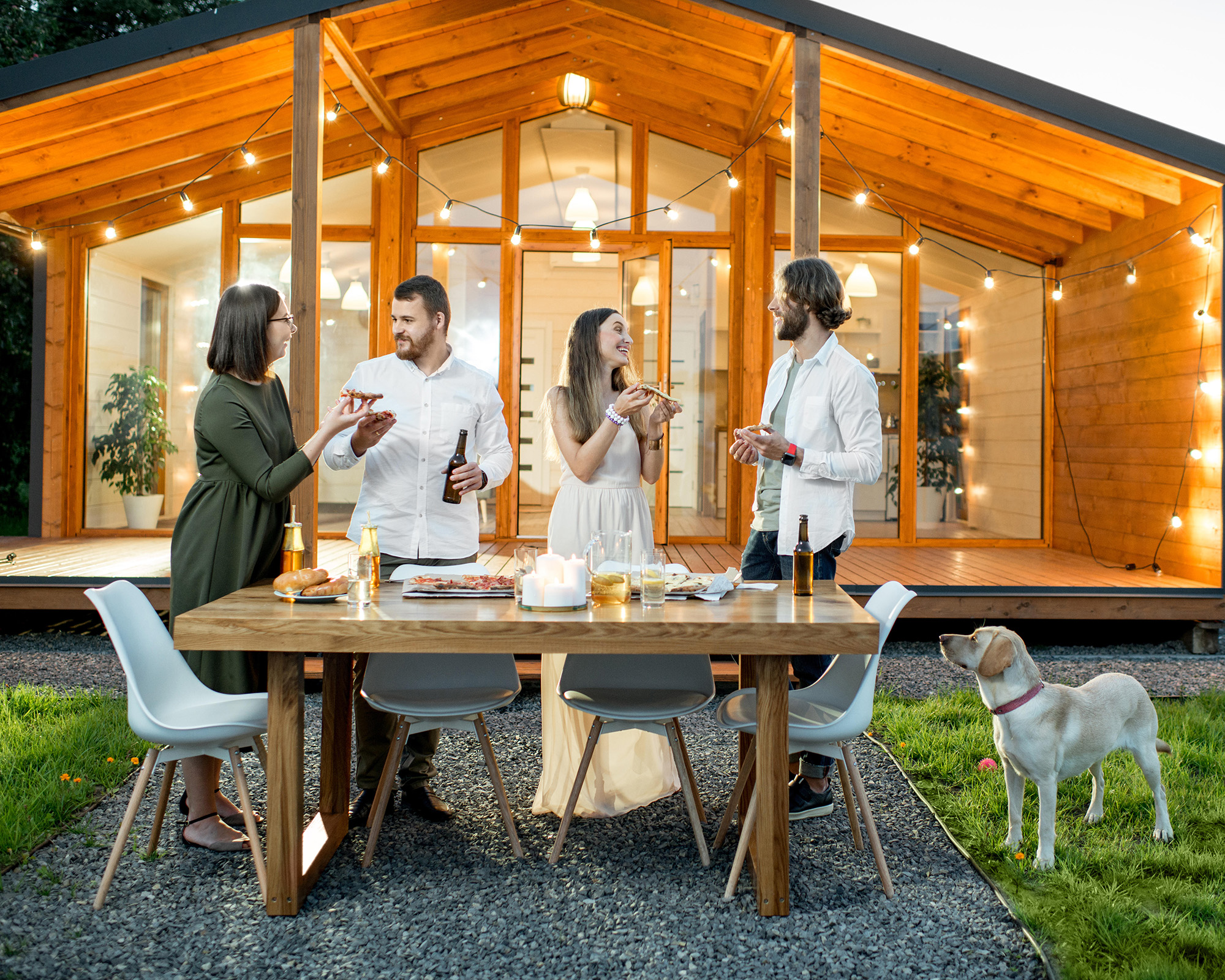 Friends having a dinner with pizza on the backyard of the modern house decorated with lights during the evening time outdoors