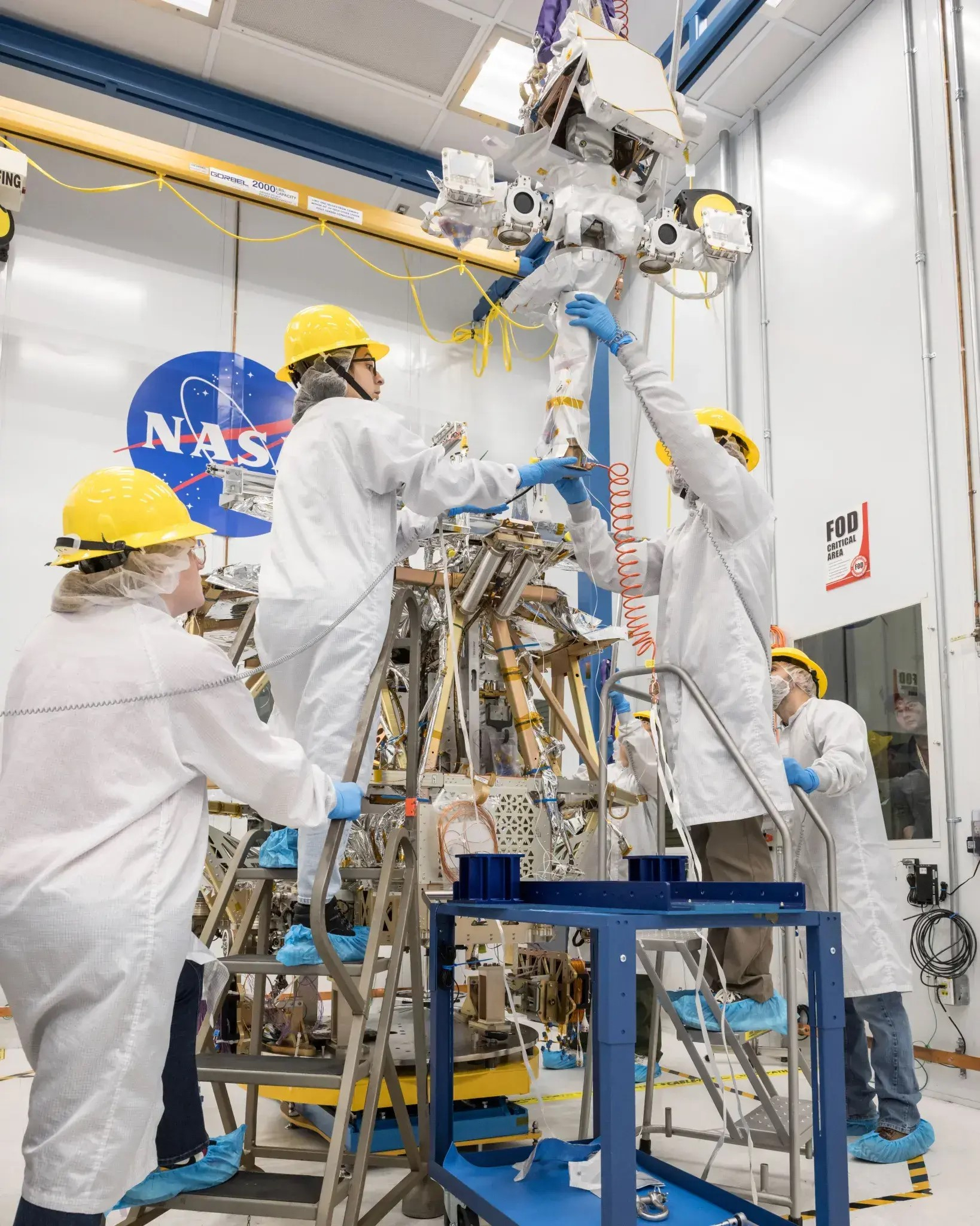 Four white-coated, yellow hard hat-wearing technicians assemble a robotic explorer in a white room.
