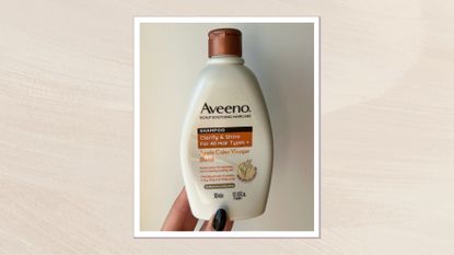 Hand holding the Aveeno Apple Cider Vinegar Shampoo bottle on a cream background