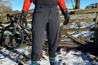The Rab Cinder Kinetic Waterproof shorts seen on a rider from the front with the fastening details on display in front of a bike leaning against a farm gate on snowy ground