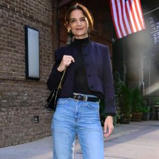 Katie Holmes stands outside on the pavement wearing blue jeans with black suede boots and a navy jacket.