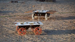 two small four-wheeled robotic rovers drive in a sandy enclosure during a test