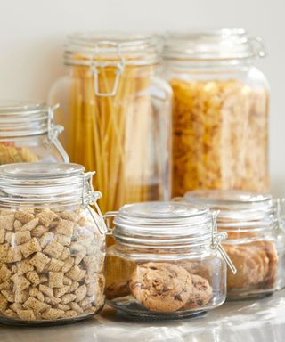 Six glass jars with pasta, cereals, and biscuits contained inside of them, on a gray granite countertop