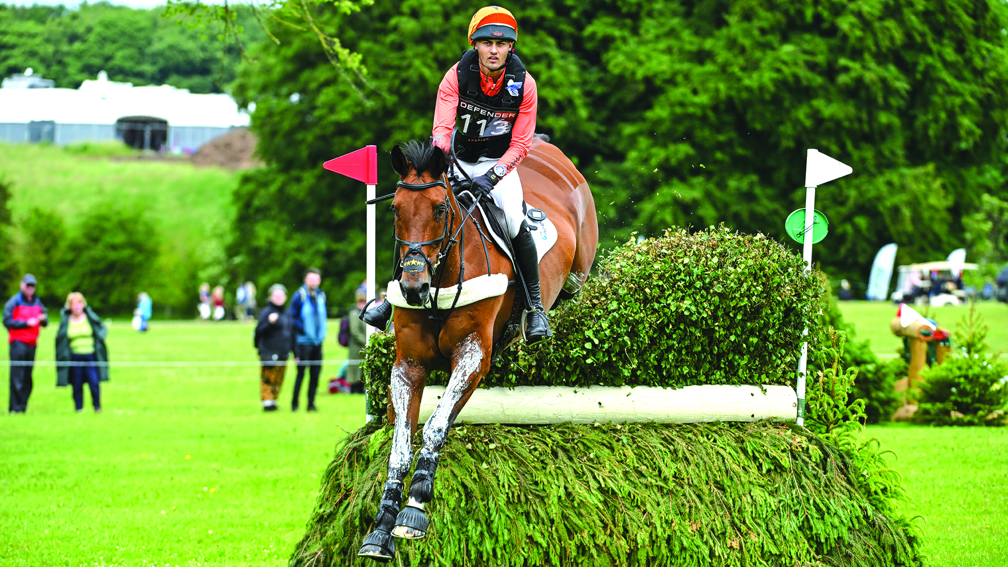 Tom Woodward, a rider in orange colours, rides bay horse Low Moor Lucky over a brush fence at Bramham Horse Trials 2025