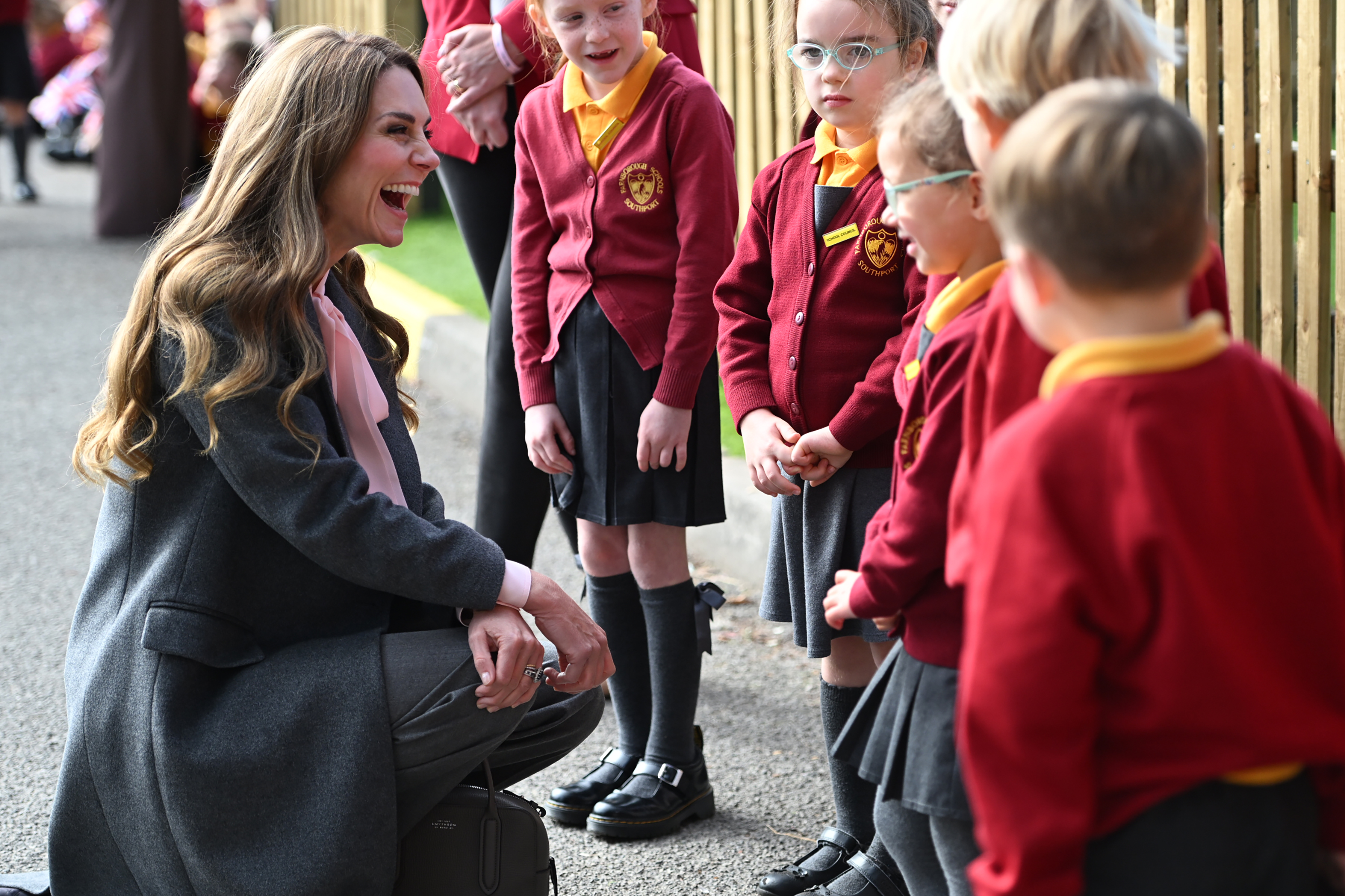 Kate Middleton wearing a gray suit leaning down and laughing with kids in red uniforms