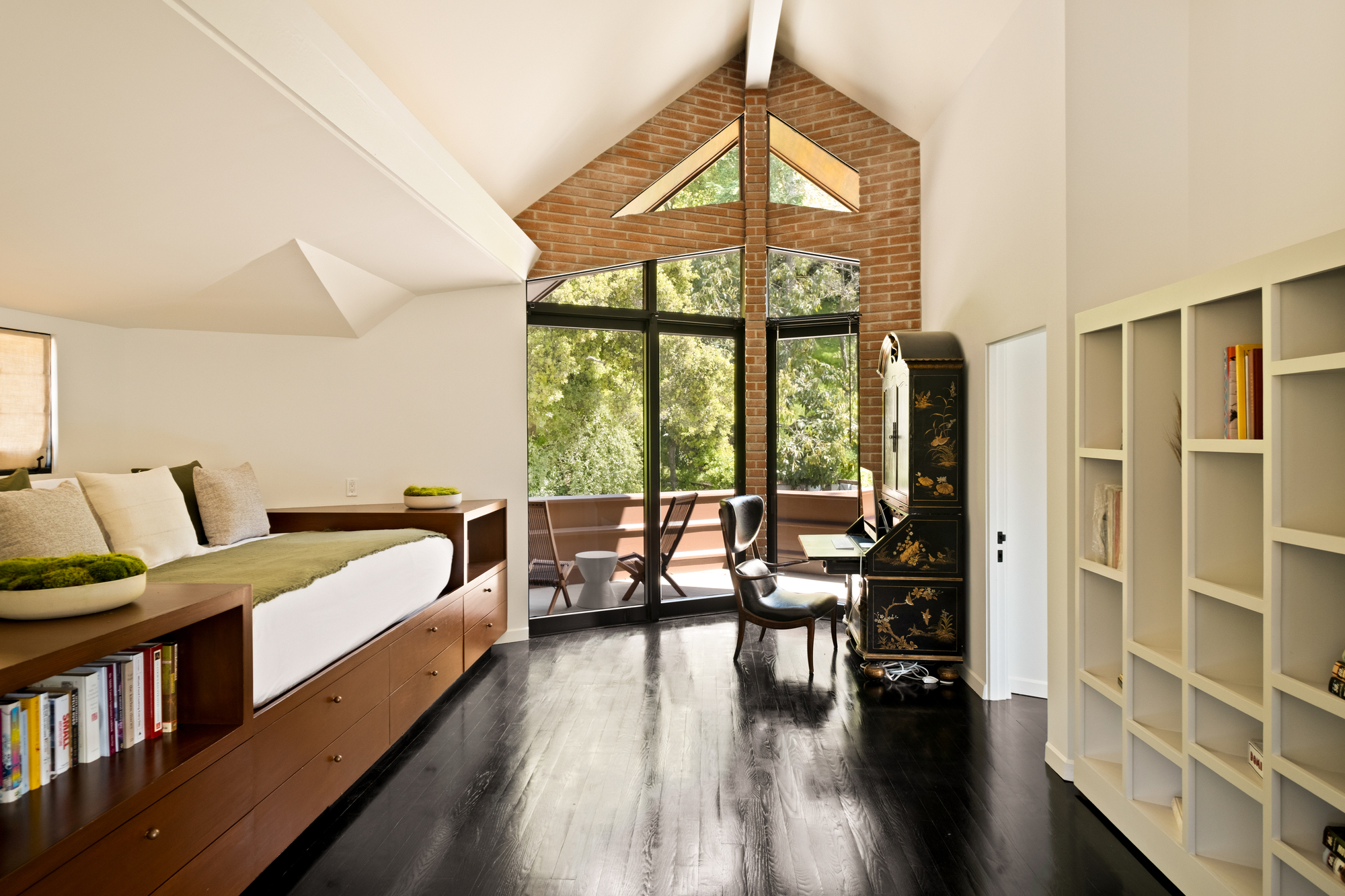 Image of a long, open, white room with black, glossy hardwood floors. There is an angular brick wall at the end with lots of windows that open up to a porch. On one side of the space is a long and low built-in daybed that also has storage.