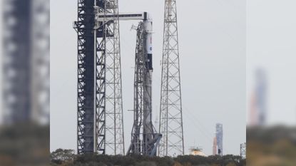 A white and black rocket is shown at a launch pad next to its launch tower and supports during the day, with a second large orange rocket with a white top and boosters pictured in the background.