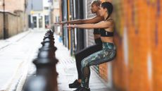 two women wearing leggings and crop top doing a wall sit in a side street with a row of pavement posts in shot