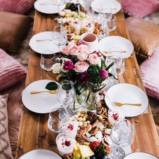 Indoor picnic featuring fresh pink roses, grazing nibbles, gold cutlery and white plates styled in beautiful tablescape on low wooden picnic table, with plethora of floor cushions.