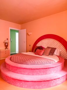 A round-shaped bed with two velvety rose steps features a semicircular bed board, matching cushions, and a peachy color palette across its walls and furniture.