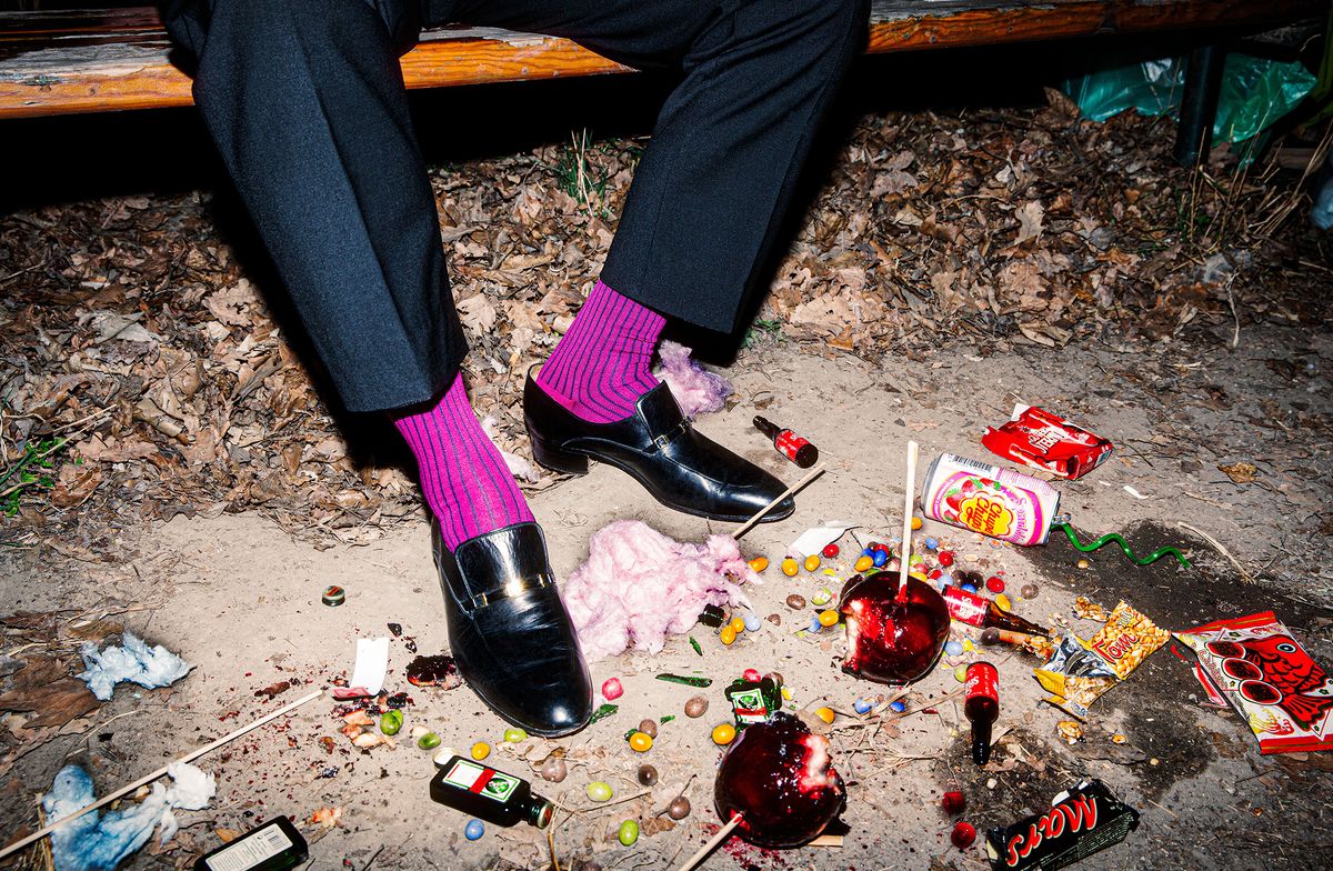 A man's legs are visible, wearing a suit and vibrant purple socks, siting on a bench, surrounded by scattered candy, discarded bottles, and trash