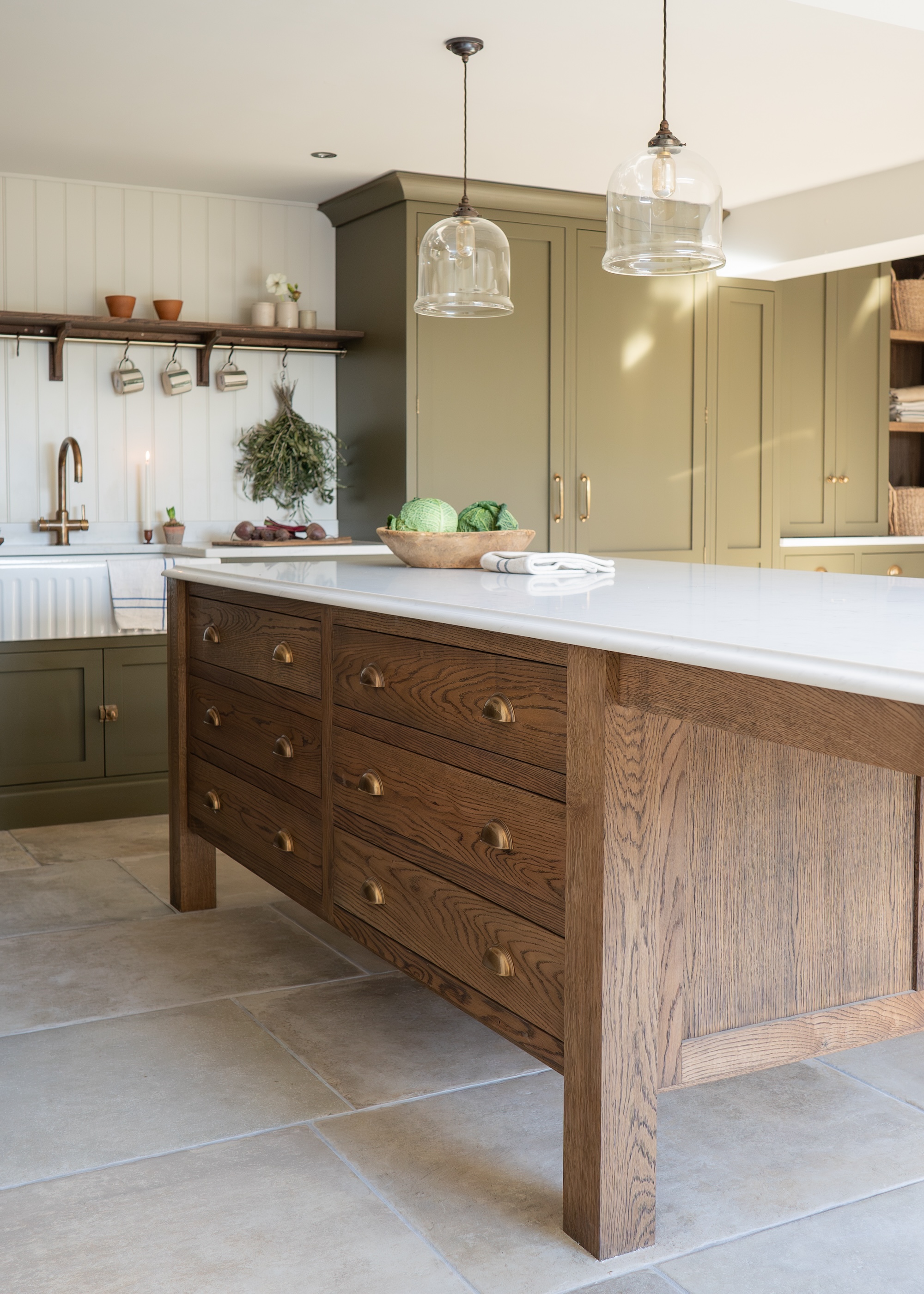 A cottage, barn style kitchen with stone flooring and a wooden island, marble worktops and sage green cupboards behind.
