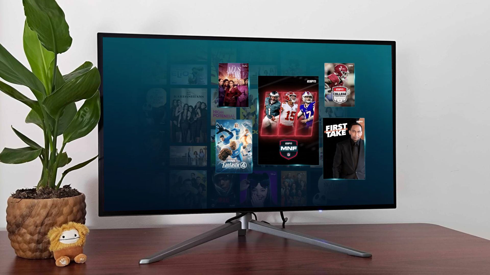 A monitor on a wooden surface with a plant and a plushie, with the screen displaying Disney Plus, Hulu, and ESPN content