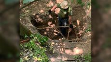 We see a man in a hat in a dirt hole and holding onto part of a rusty, decayed plane.