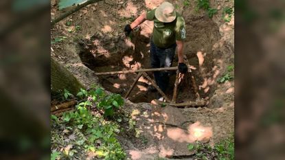 We see a man in a hat in a dirt hole and holding onto part of a rusty, decayed plane.