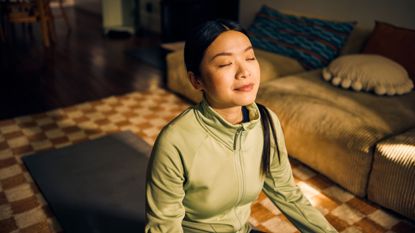 A woman in a green zipped sweater meditates with her eyes clothes. She is smiling slightly, sat on a yoga mat on the floor in a living room in front of a sofa.