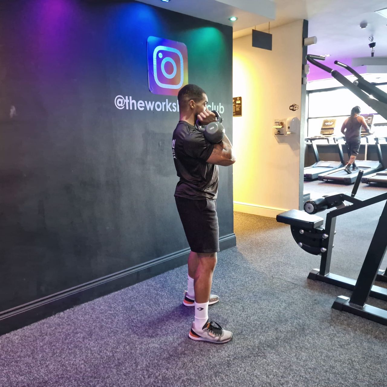 Renel Scarlett performs a kettlebell snatch in The Works Health Club gym. He is standing, feet hip-width apart, with his arms bent and a kettlebell held just under his chin. Behind him we see a person on a treadmill.