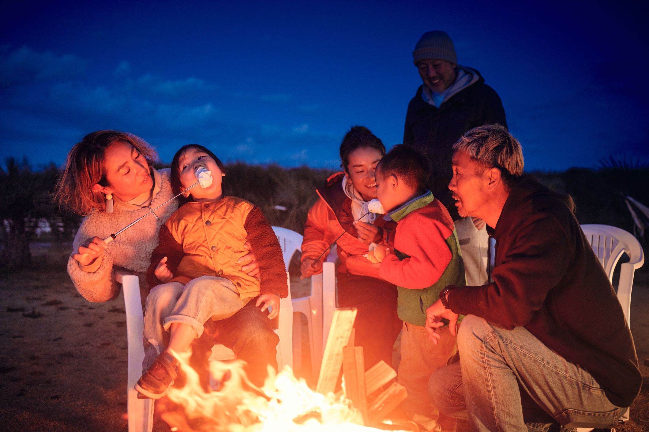 Adults help kids make smores over a campfire with plastic chairs.