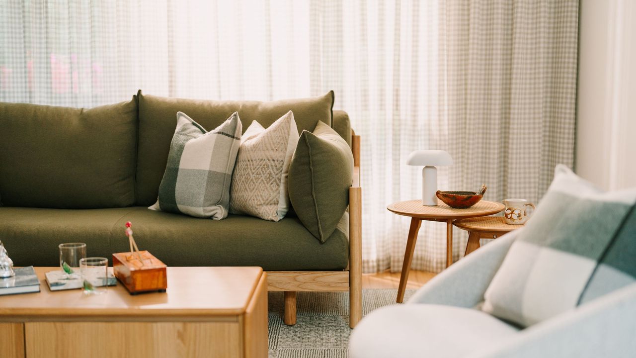 An olive green couch with cushions on top, in front of it is a coffee table with two glasses and a box. On the right side is a side table with a little chic white lamp