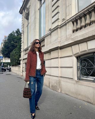 Woman wearing Chanel red tweed jacket, white t-shirt, black sunglasses, blue relaxed jeans, Chanel purse, black pointed pumps