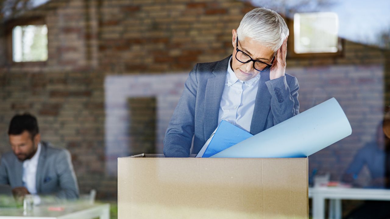 An older woman looks forlorn with a box of belongings from her office after being laid off.