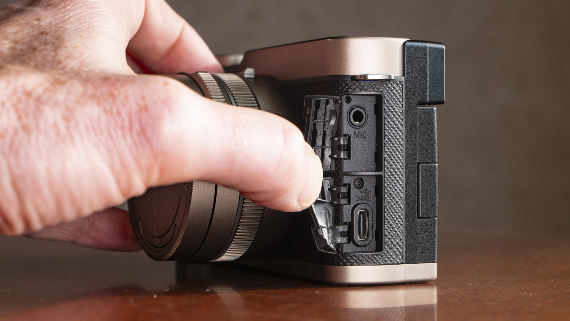 The mic and USB-C port doors opened of the Titanium Gold special edition of the Panasonic Lumix L10 compact camera, on an antique wooden table 