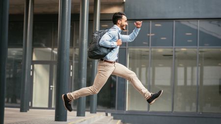 A businessman appears to fly through the air as he runs with a backpack outside an office building.