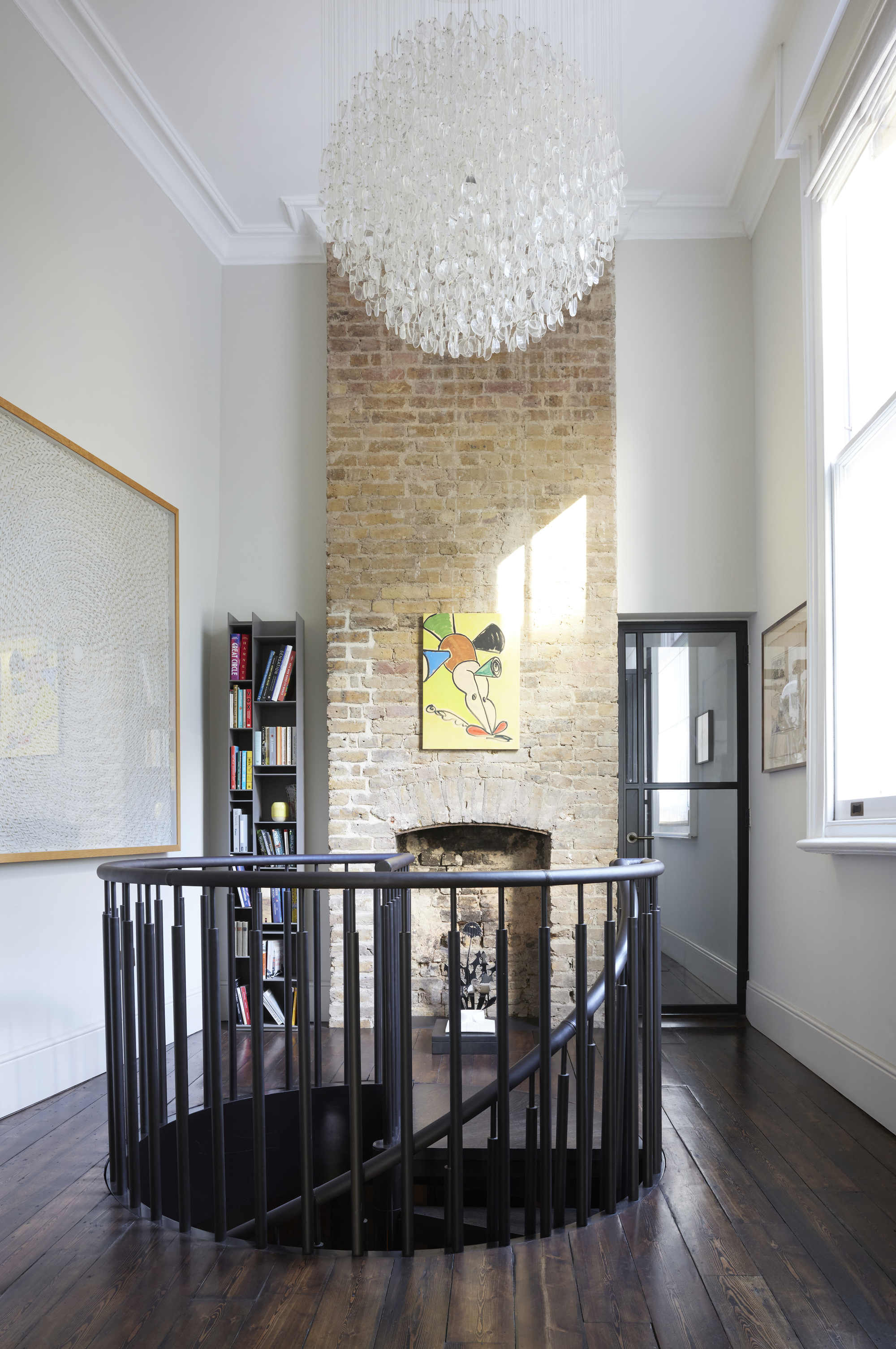 A high-ceilinged room featuring a spiral staircase with a black metal railing, a tall exposed-brick fireplace column, a large crystal globe chandelier, and dark hardwood floors.