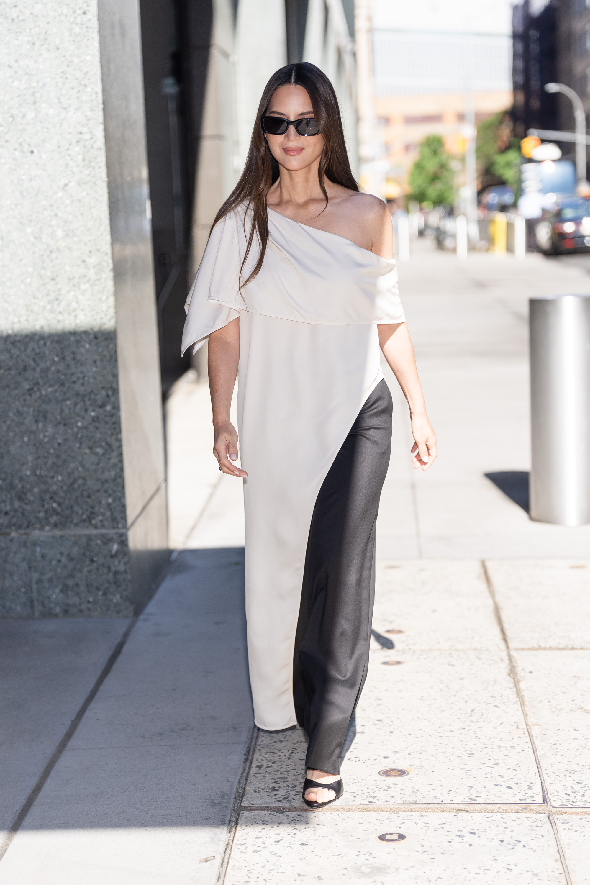 olivia munn wears a white asymmetrical dress, black trousers and black sandals.