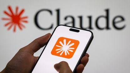 A photo of the Anthropic Claude logo on a phone in someone's hands, with the word 'Claude' and logo against a white backdrop visible in the background.
