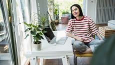Woman sits on a chair with her legs crossed underneath her, and rests one arm on a desk with a laptop on it
