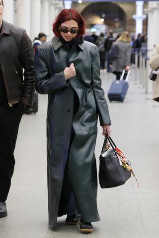 Dua Lipa wearing a long black leather coat and carrying a bag-charmed Hermès Birkin bag at St. Pancras station in London.