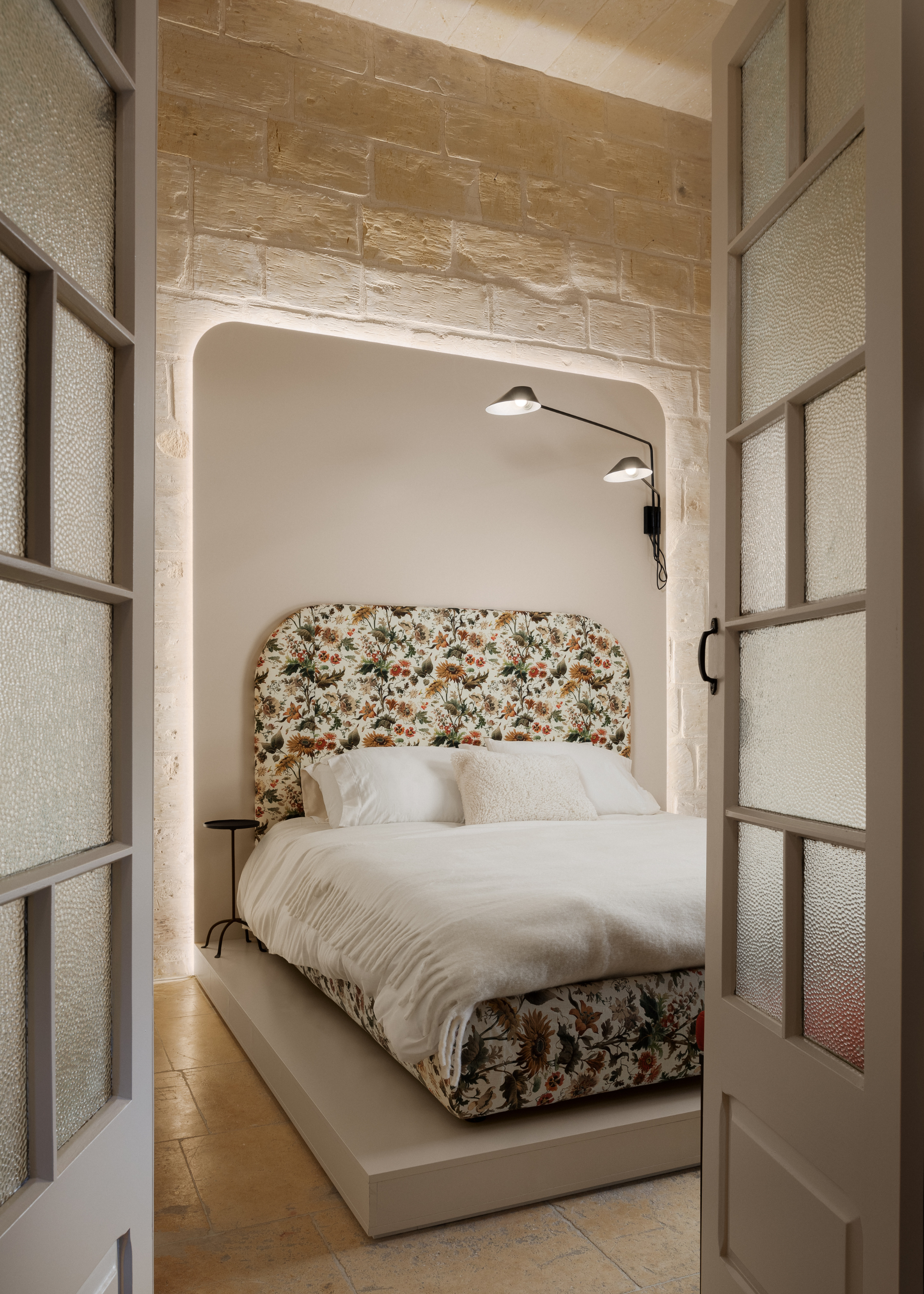 An off-white brick bedroom with an oversized backlit panel, a patterned bedstand and headboard, and a white bedscape