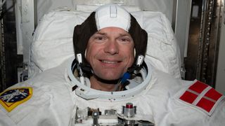 an astronaut smiles in a white spacesuit bearing the red and white flag of Denmark